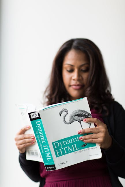 The image shows a woman with dark brown, wavy hair reading a book titled 'Dynamic HTML,' which features a stylized illustration of a flamingo on the cover. She is dressed in a black blazer over a burg